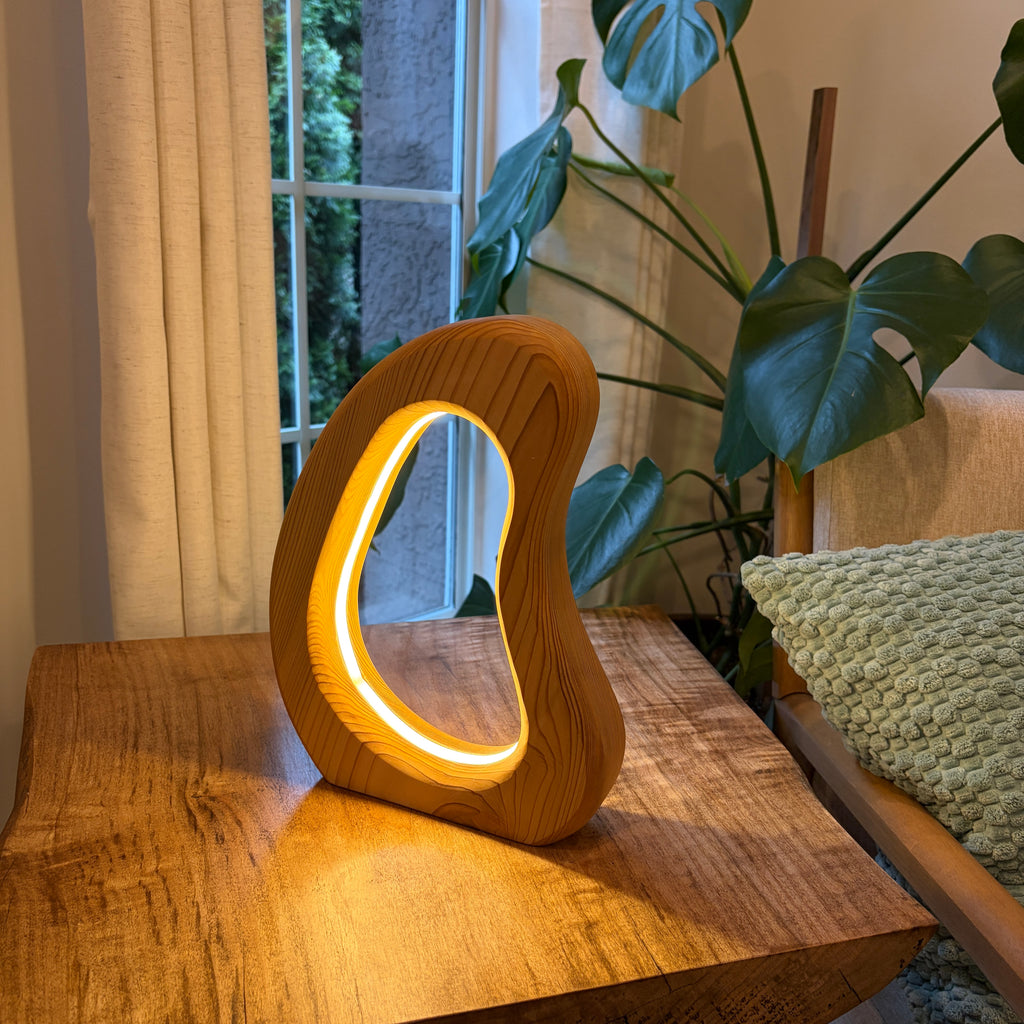 Wooden lamp with a circular design on a wooden surface next to a plant and window.