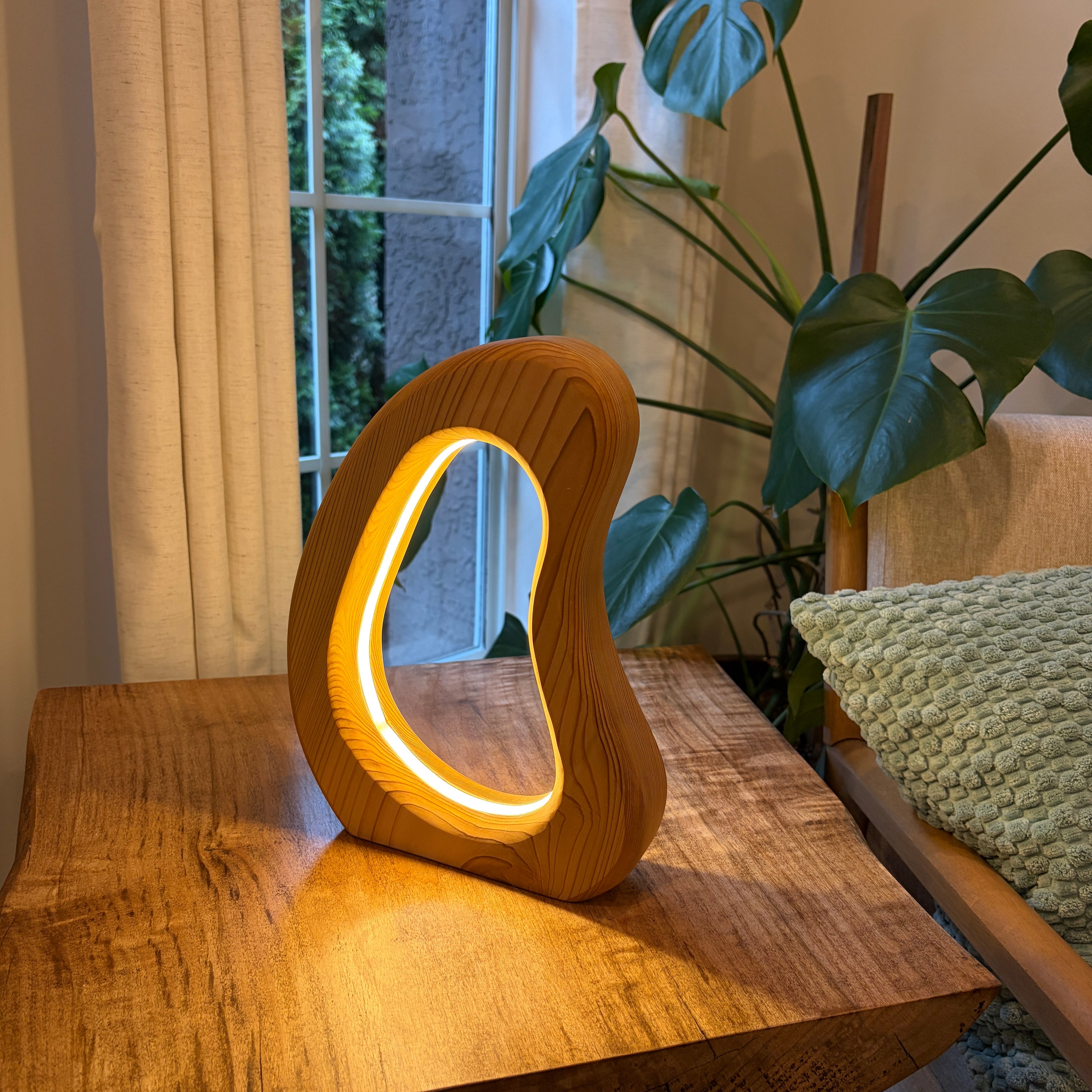 Wooden lamp with a circular design on a wooden surface next to a plant and window.