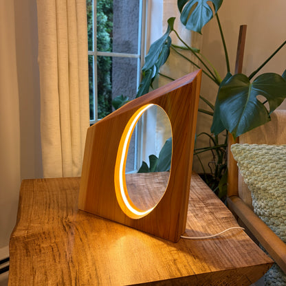 Wooden lamp with circular light on a wooden surface near a window with plants in the background