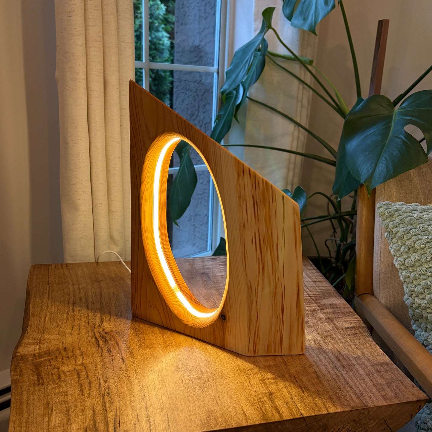 Wooden lamp with circular light on a wooden surface, surrounded by plants and a window.